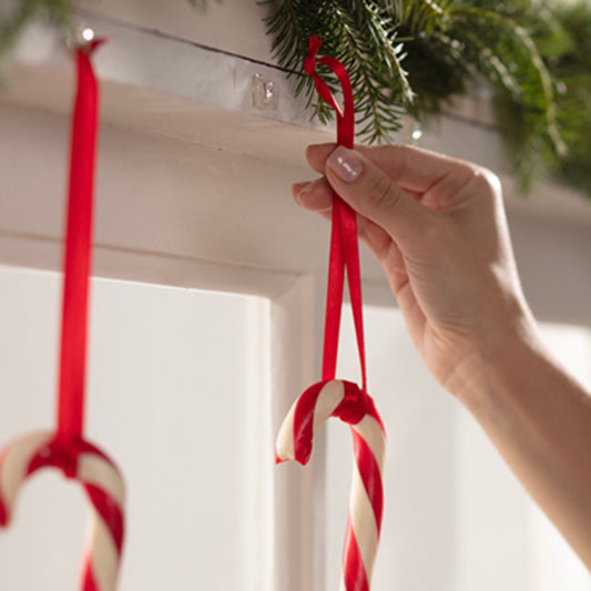 Eine Hand hängt eine rot-weiße Zuckerstange mit einem roten Band an einen transparenten Command™ Mini-Haken (17006CLRFGN) der 3M Deutschland GmbH, der unter Grünzeug auf einem weißen Kaminsims angebracht ist. Eine weitere Zuckerstange hängt bereits daneben.