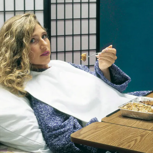 Eine Person mit lockigem Haar liegt im Bett an einem Kissen gelehnt, trägt einen blauen Pullover und einen Servocare Kleiderschutz für Erwachsene von Servoprax GmbH. Sie isst mit einer Gabel Essen aus einem Folienbehälter. Vor ihnen steht ein Holztisch und im Hintergrund ein Shoji-Paravent.