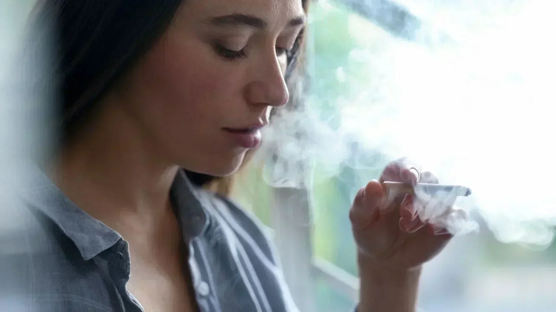 Eine Frau sitzt in der Nähe eines Fensters und raucht eine Zigarette. Der Rauch steigt in die Luft, während sie nachdenklich in die Ferne blickt. Sie trägt ein graues Hemd und hat braune Haare, die leicht im Licht glänzen.