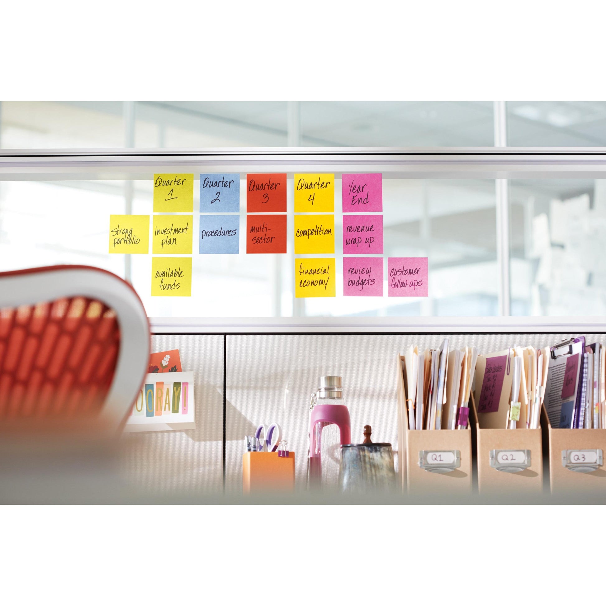 Eine Reihe von 3M Deutschland GmbH Post-it® Super Sticky Notes, 76 mm x 76 mm, 90 Blatt/Block, 100% PEFC, ist auf einer Glastrennwand im Büro über organisierten Vorräten, Ordnern und Büchern mit einem orangefarbenen Stuhl davor angeordnet.