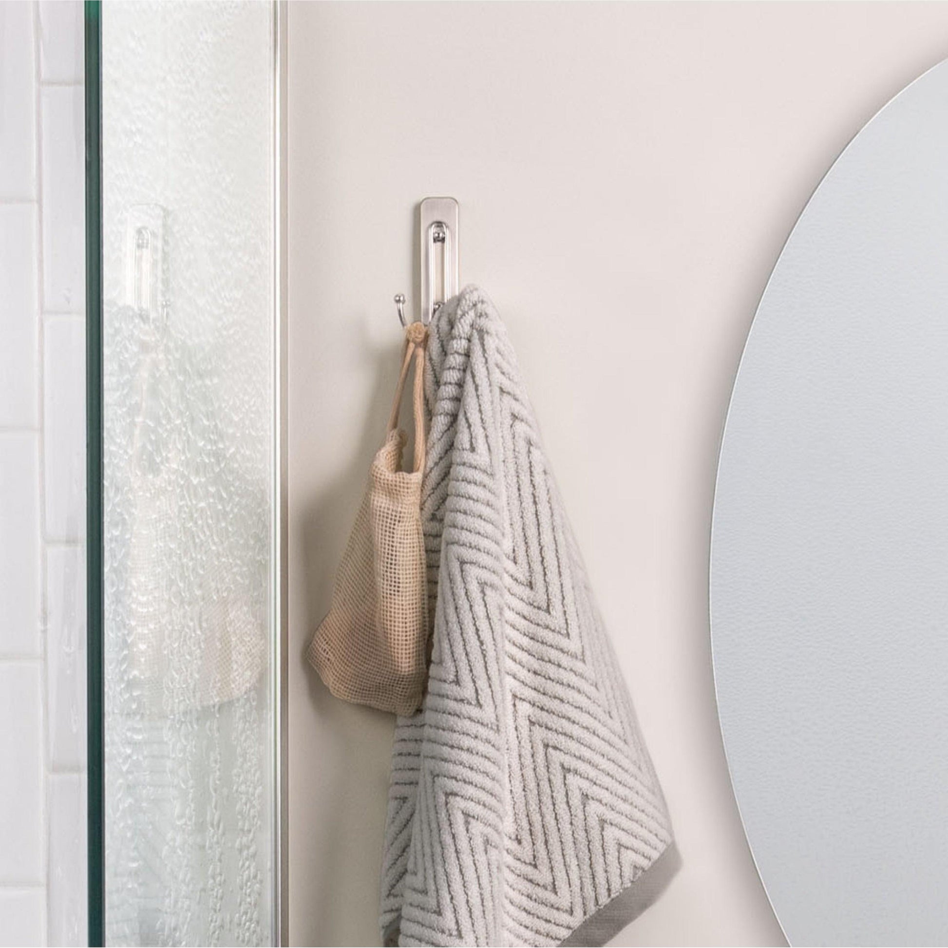 Ein gemustertes graues Handtuch und eine beigefarbene Netztasche hängen an einem satinvernickelten Command™ Großer Badezimmer-Doppelhaken von 3M Deutschland GmbH neben einem runden Spiegel und einer strukturierten Glastür im Badezimmer.
