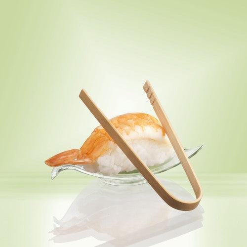 Une paire de pinces à fingerfood de 10 cm PAPSTAR en bambou de PAPSTAR GmbH repose sur un plat en verre à côté de nigiri aux crevettes - idéal pour servir des snacks ou des fingerfoods de fête - sur un fond avec un dégradé de couleur vert pâle.