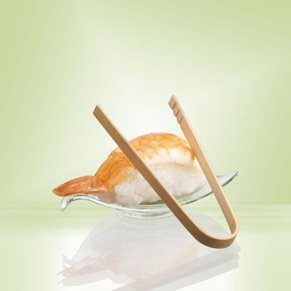 Une paire de pinces à fingerfood de 10 cm PAPSTAR en bambou de PAPSTAR GmbH repose sur un plat en verre à côté de nigiri aux crevettes - idéal pour servir des snacks ou des fingerfoods de fête - sur un fond avec un dégradé de couleur vert pâle.