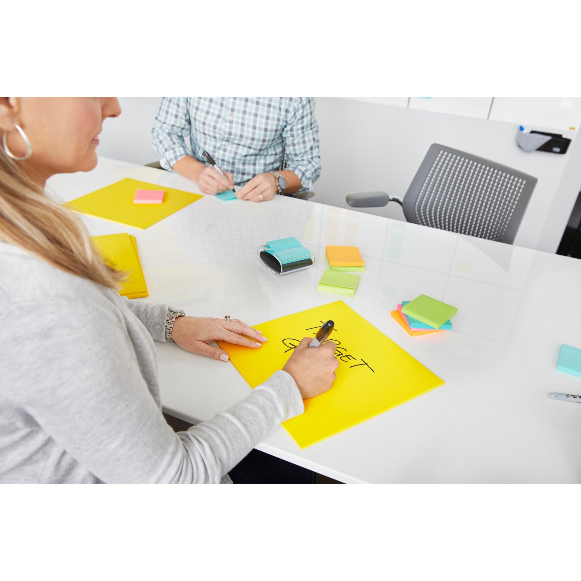 Zwei Personen sitzen an einem weißen Tisch, der mit bunten Haftnotizen bedeckt ist. Eine schreibt "TARGET GROUP" auf einen Post-it® Super Sticky Big Note (BN11-EU, Gelb, 279x279 mm) von 3M Deutschland GmbH; weitere Zettel und Zubehör liegen in der Nähe.