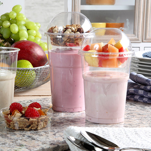 Twee yoghurtparfaits in plastic bekers, elk afgesloten met een Starpak 50 Dom-deksel „Hurricane“ PET rond glashelder Ø 9,5 cm, staan op een keukentoonbank - de ene met muesli en noten, de andere met fruit. Op de achtergrond zijn verse bessen en gestapelde borden te zien.