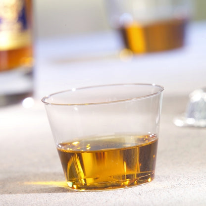 Een Starpak 15 drinkbeker, PS 0,1 l glashelder, bevat een kleine hoeveelheid amberkleurige vloeistof op een lichte ondergrond, terwijl op de achtergrond een andere beker en een fles onscherp zijn.