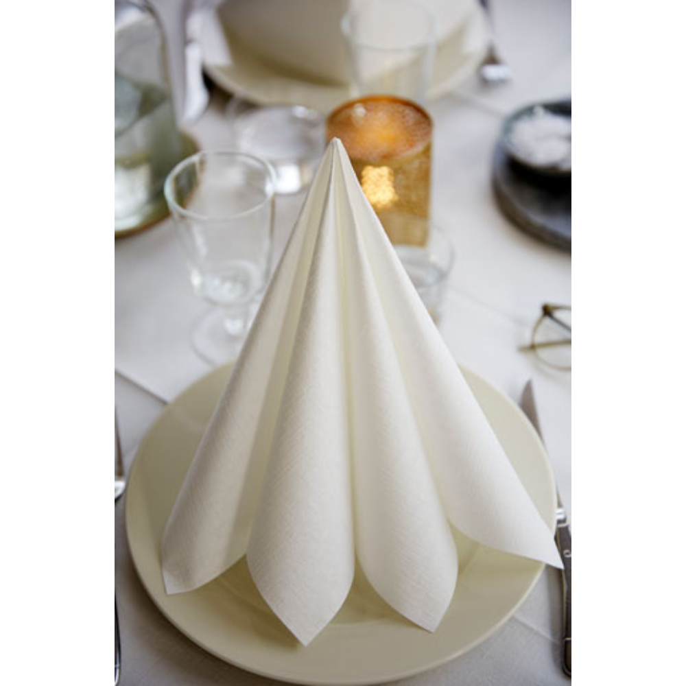 Une serviette Dunilin® 40 x 40 cm de Duni GmbH pliée proprement se tient debout sur une assiette blanche, entourée de verres et de couverts sur une table dressée. Une bougie allumée et quelques verres rehaussent l'ambiance avec une décoration de table durable en arrière-plan.