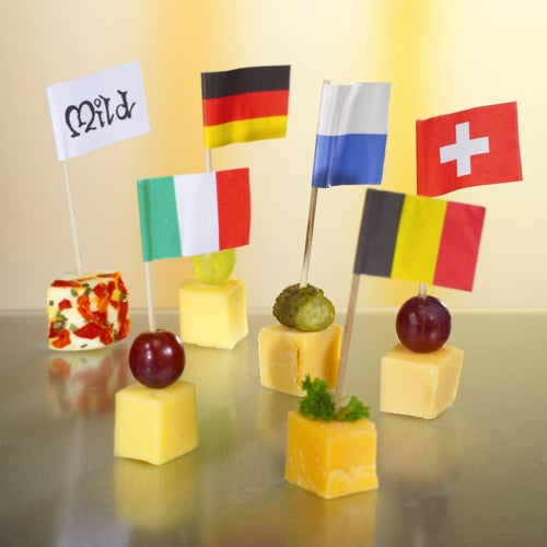 Six cubes de fromage sont garnis de raisins ou d'herbes et décorés pour une présentation élégante et écologique avec les petits drapeaux de décoration blancs de 8 cm de PAPSTAR GmbH, portant les inscriptions « Mild », Allemagne, Italie, Finlande, Suisse ou Belgique sur chaque drapeau.