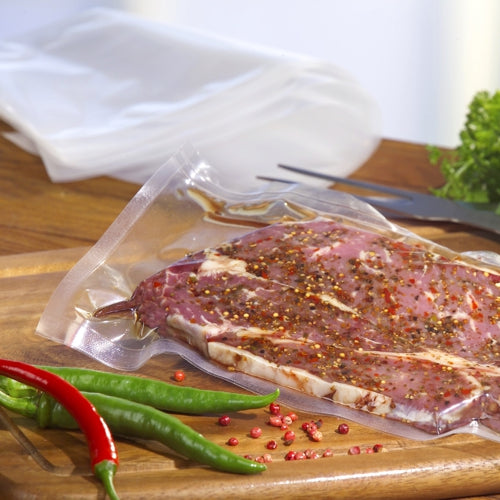 Un sac Starpak 100 à bord scellé, PA/PE transparent, rempli de viande marinée repose sur une planche à découper en bois au milieu de piments et de grains de poivre rose. Des légumes-feuilles et des ustensiles complètent la scène - idéal pour une utilisation avec une machine à vide à cloche.