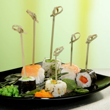 Une assiette noire avec différents sushis, chaque pièce piquée avec PAPSTAR 40 pics à fingerfood « pure » de PAPSTAR GmbH et garnie d'herbes et d'œufs de poisson, est élégamment présentée devant un fond avec un dégradé de vert.