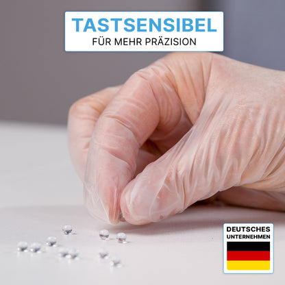 Eine Hand in ARNOMED VINYL PF Tastsensible Einmalhandschuhe von ARNOWA GmbH greift eine silberne Kugel von mehreren auf weißer Fläche. Oben steht: "Tastsensibel für mehr Präzision." Unten rechts ist eine Deutschland-Flagge zu sehen.