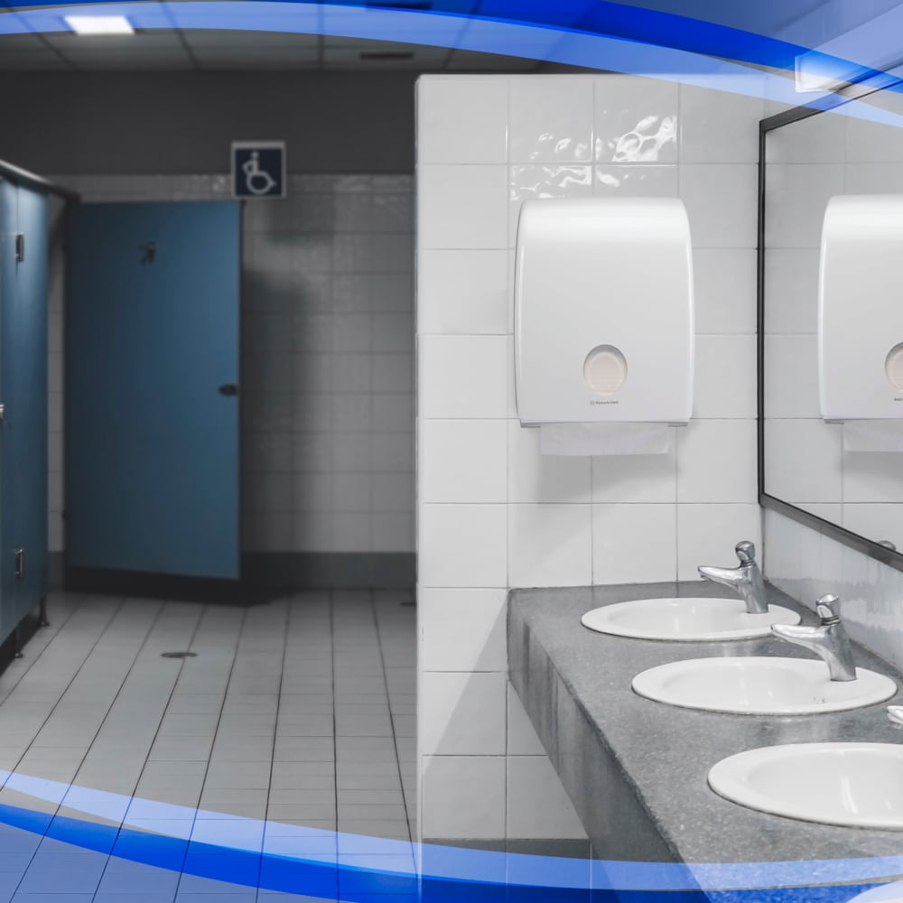 Une toilette publique avec trois lavabos, un grand miroir, des portes de cabine bleues - dont une accessible en fauteuil roulant - et des sols et murs carrelés blancs dispose d'un distributeur Aquarius™ pour serviettes pliées de Kimberly-Clark GmbH au-dessus des lavabos.