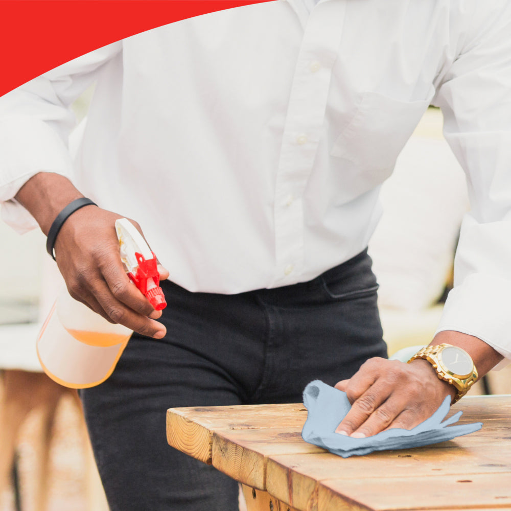 Eine Person mit weißem Hemd und Uhr reinigt einen Holztisch mit einem blauen Tuch, möglicherweise WypAll® L20 General Clean - BRAG™ Box von Kimberly-Clark GmbH. Das Bild hat oben einen roten geschwungenen Rand, der die Liebe zum Detail betont.