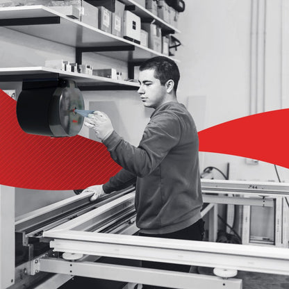 In een werkplaats stelt een persoon een machinedeel in naast een werkbank die is uitgerust met de WypAll® L10 papieren doek centrefeed voor Reach+™ (38,00 x 18,30 cm) van Kimberly-Clark GmbH voor snelle reiniging in de detailhandel; op de achtergrond zijn planken en een rode grafiek te zien.