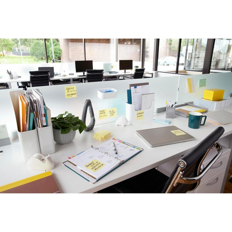 Un bureau moderne avec un ordinateur portable, un planificateur, une tasse de café, une plante, des fournitures de bureau et des notes Post-it® jaunes 3M (76x76 mm, 100 feuilles/bloc), idéal pour des notes rapides. Les dossiers sont organisés et de grandes fenêtres offrent une vue sur l'extérieur; les bureaux sont alignés en rangée.