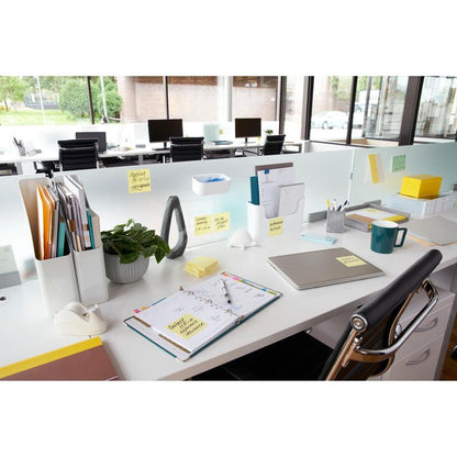 Un bureau moderne avec un ordinateur portable, un planificateur, une tasse de café, une plante, des fournitures de bureau et des notes Post-it® jaunes 3M (76x76 mm, 100 feuilles/bloc), idéal pour des notes rapides. Les dossiers sont organisés et de grandes fenêtres offrent une vue sur l'extérieur; les bureaux sont alignés en rangée.