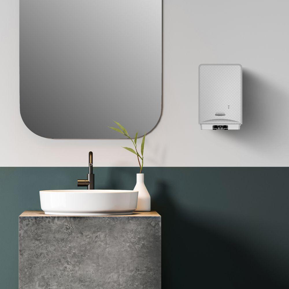 Een moderne badkamer beschikt over een ronde spiegel, een witte wastafel, een zwarte kraan en de Scott® Control™ Schuim-handreiniger (4 patronen) van Kimberly-Clark GmbH voor optimale handhygiëne. Naast de wastafel staat een vaas met groene bladeren, rechts daarvan bevindt zich een boiler.