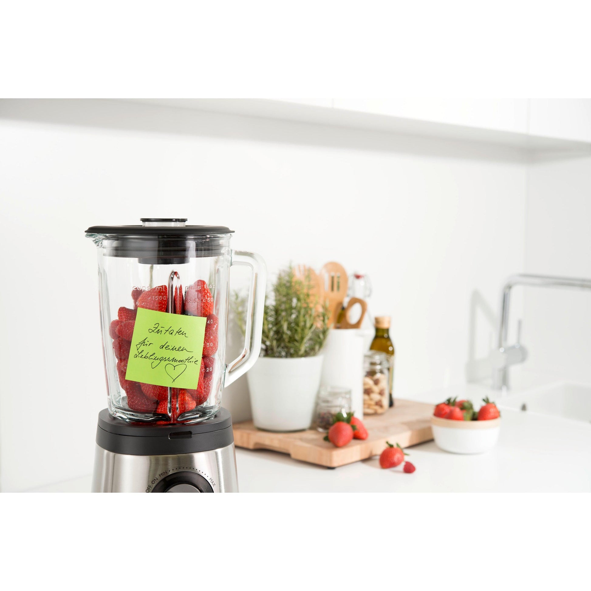 Ein Mixer mit Erdbeeren steht auf einer weißen Küchenzeile. Ein Post-it® Super Sticky Note (76 x 76 mm, 90 Blatt, 100 % PEFC) der 3M Deutschland GmbH ist an dem Behälter befestigt. In der Nähe befinden sich Utensilien, Topfpflanzen, Olivenöl und weitere Erdbeeren auf einem Schneidebrett.