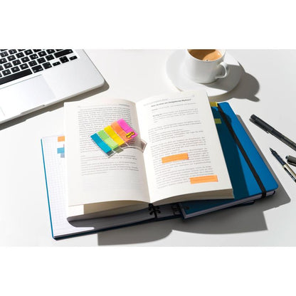 Sur un bureau bien éclairé se trouve un livre ouvert, étiqueté avec des Post-it® Index Signets Mini 3M Deutschland GmbH (11,9 x 43,2 mm, 100/distributeur), au-dessus d'un cahier bleu et d'un ordinateur portable, à côté de stylos et de café sur la surface blanche.