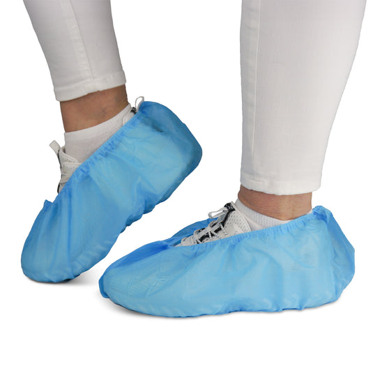 Une personne porte un pantalon blanc et des baskets recouvertes de surchaussures ARNOMED ABS de la société ARNOWA GmbH. Ces surchaussures jetables antidérapantes (sachet de 100 pièces) protègent les chaussures et les sols ; elles sont idéales pour les environnements où l'hygiène est primordiale.