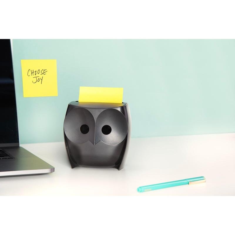 Een zwarte Post-it® Z-Notes dispenser "Uil" (3M Deutschland GmbH) met gele Super Sticky Z-Notes staat op een wit bureau, daarnaast een laptop en een blauwe pen. Aan de muur hangt een "CHOOSE JOY"-notitie tegen een lichtblauwe achtergrond.