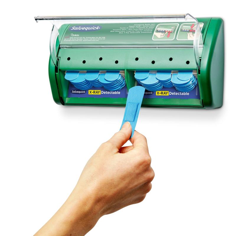 Een hand trekt een verband uit een groene Salvequick röntgendetecteerbare pleisterdispenser (Orkla Wound Care AB), die 2x30 pleistervullingen bevat, waarbij extra verbanden zichtbaar zijn binnenin de dispenser.