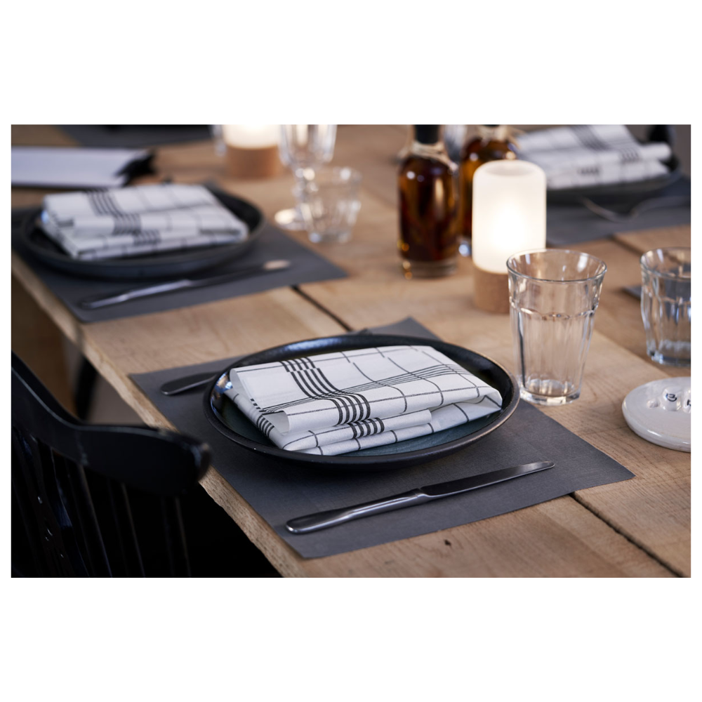 Een eetset bestaat uit borden in Linnea Black van Duni GmbH en geruite servetten. Op de achtergrond staan drie glazen flessen naast brandende kaarsen. Waterglazen en bestek worden elegant gepresenteerd op een Duni Dunicel®-placemat 30 x 40 cm, waarbij de lichte houten tafel de eenvoudige elegantie benadrukt.