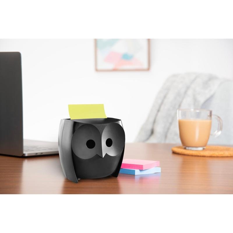 Een 3M Deutschland GmbH Post-it® Z-Notes Dispenser, Uil, Zwart houdt gele notities vast, daarnaast liggen roze en blauwe Super Sticky Z-Notes (45 vel/blok, 2 blokken/pak) op een bureau met een laptop, een koffiekopje en wandkunst op de achtergrond.