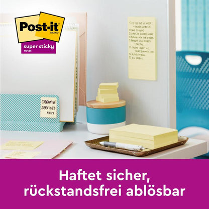 Sur un bureau se trouvent des Post-it® Super Sticky 100 % Notes de Recyclage (47,6 x 47,6 mm, 70 feuilles/bloc, 100 % PEFC) de 3M Deutschland GmbH, avec un stylo, des dossiers et une note sur le séparateur. Le logo apparaît; le texte allemand dit : "Colle en toute sécurité, se détache sans résidu."