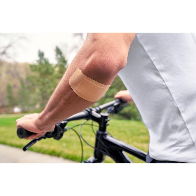 Een persoon die een wit shirt draagt en buiten fietst, heeft een Nexcare™ Ultra Stretch MAXI Comfort Flexibele Pleister (50 mm x 101 mm) van 3M Deutschland GmbH op de blote bovenarm; op de achtergrond zijn groen gras en bomen te zien.