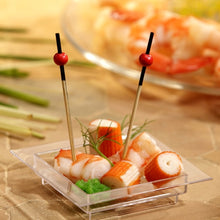 Sur un plat carré transparent se trouvent du surimi, des crevettes, une garniture verte et deux PAPSTAR 250 piqueurs Fingerfood de la PAPSTAR GmbH avec des perles rouges. En arrière-plan, des crevettes floues sont visibles sur une assiette.