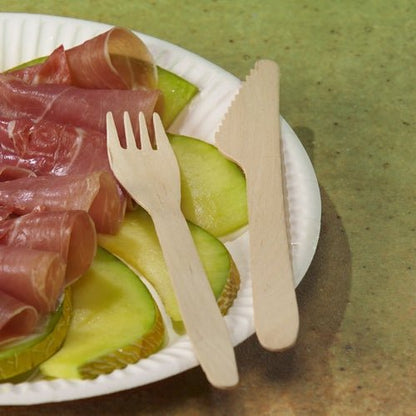 Sur une surface tachetée de vert, des tranches de pomme verte et du prosciutto plié reposent sur une assiette blanche, accompagnés de PAPSTAR fourchettes en bois "pure" 16,5 cm - écologiques et stables de PAPSTAR GmbH (25 pièces).