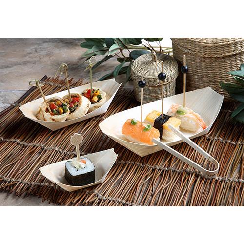 Fingerfood-sushi en voorgerechten worden geserveerd in PAPSTAR 50 Fingerfood-schalen, hout „pure“ „bootjes“ van de PAPSTAR GmbH, elk stuk op een decoratieve prikker en gepresenteerd op bamboe-placemats met groene bladeren en gevlochten manden op de achtergrond.