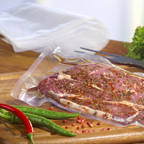 De la viande crue et épicée est mise sous vide dans un PAPSTAR sachet à souder 100 transparent (PA/PE) de la PAPSTAR GmbH sur une planche à découper en bois, entourée de piments, de grains de poivre, de couverts et de légumes-feuilles.
