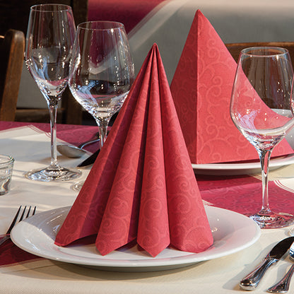 Une serviette pliée PAPSTAR 50 serviettes "ROYAL Collection" 1/4 pli 40 x 40 cm rouge de PAPSTAR GmbH est dressée sur une assiette blanche, des verres à vin, des couverts et une nappe à motifs rouges complètent la décoration de table élégante.