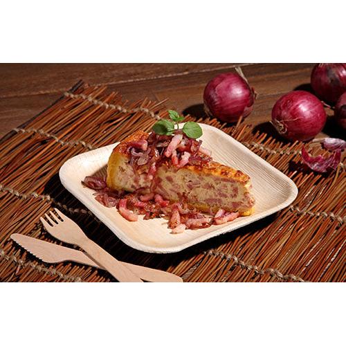 Une tranche de tortilla espagnole avec des oignons et des herbes est servie sur une assiette carrée PAPSTAR 25, feuille de palmier « pure » de PAPSTAR GmbH, posée sur un tapis tressé avec des couverts en bois et des oignons rouges en arrière-plan.