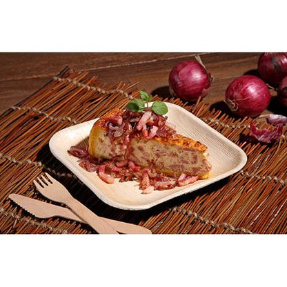 Une tranche de tortilla espagnole avec des oignons et des herbes est servie sur une assiette carrée PAPSTAR 25, feuille de palmier « pure » de PAPSTAR GmbH, posée sur un tapis tressé avec des couverts en bois et des oignons rouges en arrière-plan.