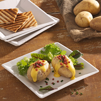 Deux pommes de terre cuites au four avec une sauce au fromage jaune, des morceaux de bacon et de la ciboulette sont servies sur une assiette en carton carrée de 22,5 cm au design "pure" de PAPSTAR GmbH. Des légumes-feuilles accompagnent le plat, avec du pain grillé et des pommes de terre entières en arrière-plan.