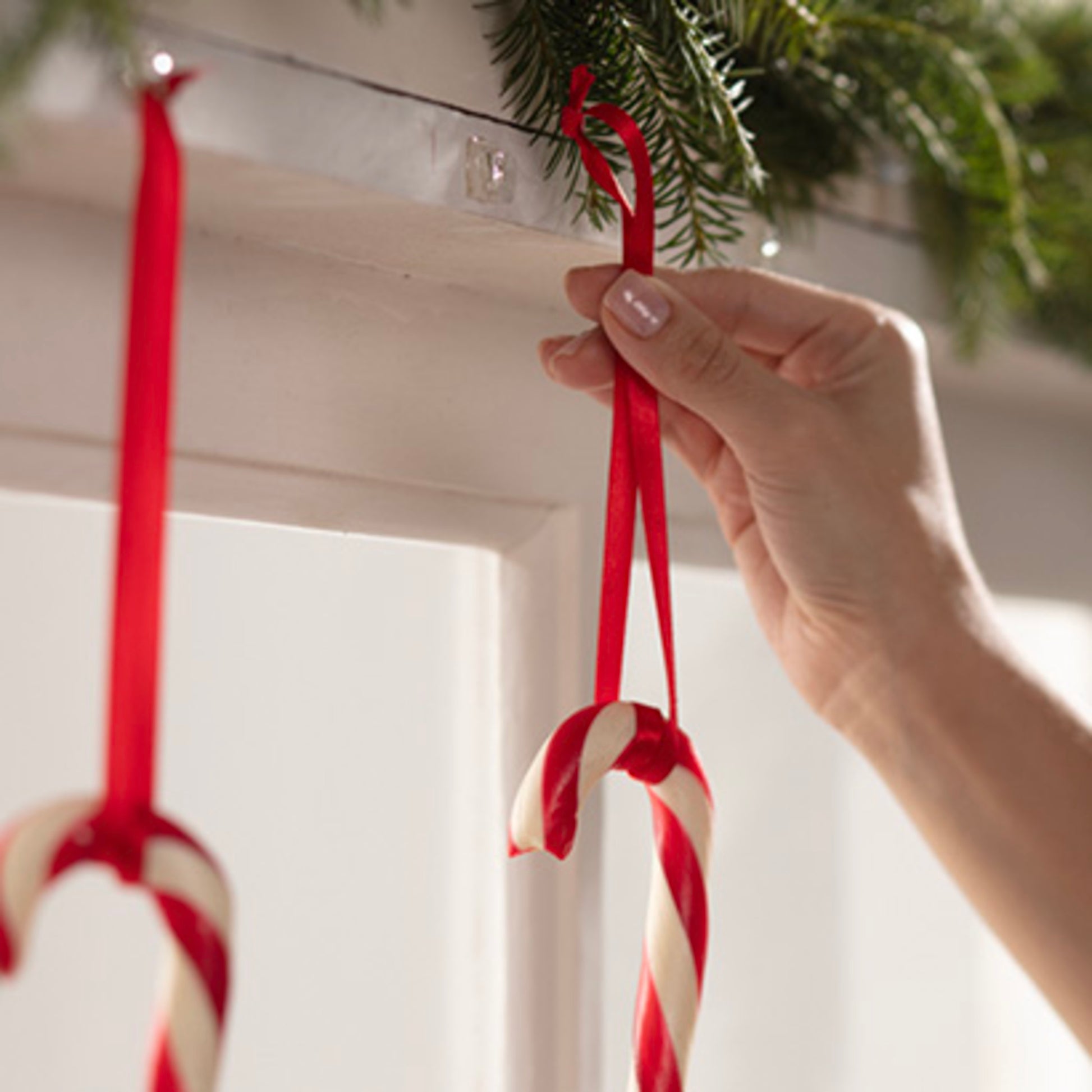 Eine Hand hängt eine rot-weiße Zuckerstange mit einem roten Band an einen transparenten Command™ Mini-Haken (17006CLRFGN) der 3M Deutschland GmbH, der unter Grünzeug auf einem weißen Kaminsims angebracht ist. Eine weitere Zuckerstange hängt bereits daneben.