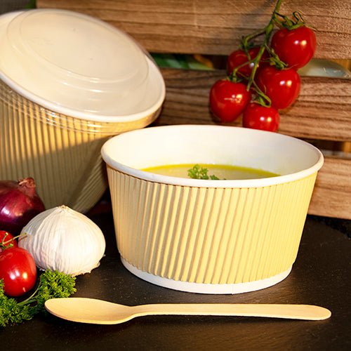 Een bruin/witte PAPSTAR ronde soepbeker, 11 cm Ripple Wall (25 stuks) van PAPSTAR GmbH is gevuld met gele soep, naast een houten lepel, knoflook, ui, cherrytomaten aan de wijnstok, peterselie en een andere gesloten schaal op de achtergrond.