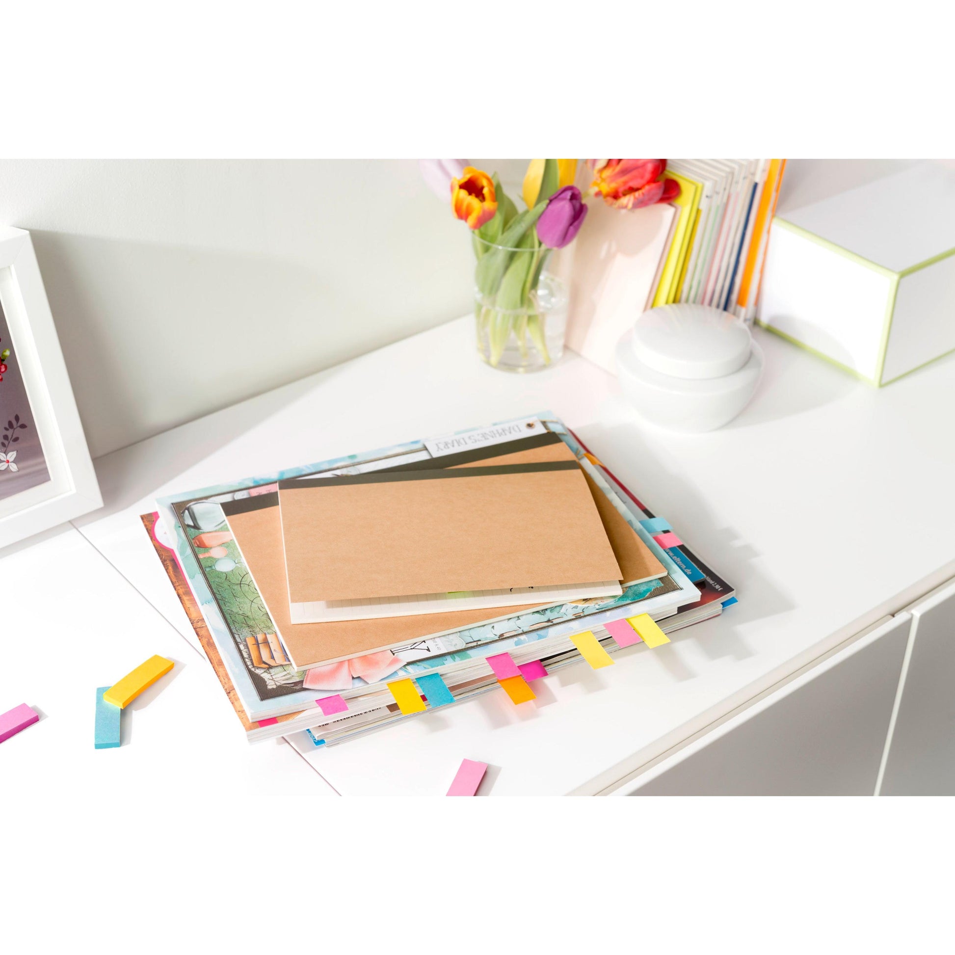 Auf einem weißen Schreibtisch liegt der Post-it® Page Marker (15 mm x 50 mm, 50 Blatt/Block, 100% PEFC) der 3M Deutschland GmbH zwischen gestapelten Büchern und Notizbüchern mit bunten Markern, verstreuten Notizen, einer Glasvase mit Blumen und Schreibwaren.