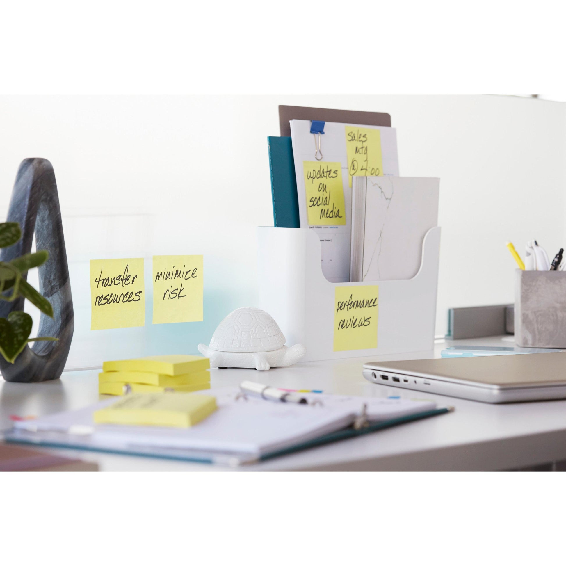 Ein moderner Bürotisch mit einem 3M Post-it® Super Sticky Notes Block (76 mm x 76 mm, 90 Blatt, 100% PEFC) auf einem Glastrenner, einer Schildkrötenfigur, Ordnern in einem Dokumentenhalter, einem geschlossenen Laptop, Stiften und einem Notizbuch.