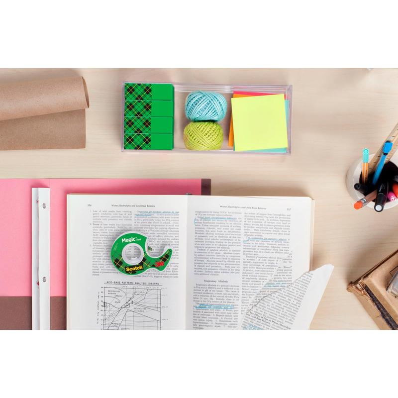 Een werkruimte met een open boek, millimeterpapier, gekleurde garenballen, plaknotities, pennen in een beker, gum, een bruine rol pakpapier en een rol 3M Deutschland GmbH Scotch® Magic™ Onzichtbare Tape op een lichte houten tafel.