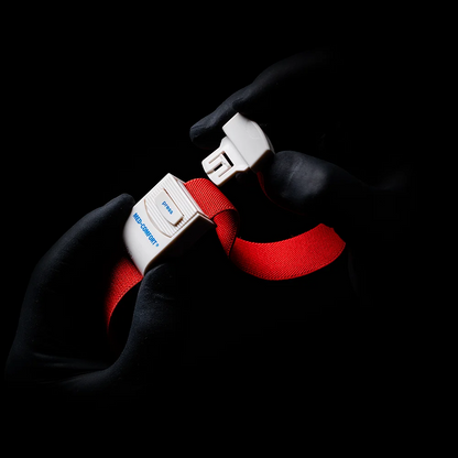 Des mains gantées, rappelant un garrot AMPri MED-COMFORT, saisissent la boucle d'une ceinture de sécurité rouge vif devant un fond ombragé. Sur la boucle, le mot « Blue » et la lettre « B » sont visibles.