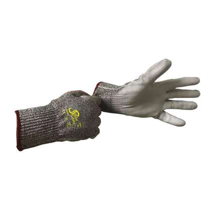 Une personne porte un gant de protection contre les coupures AMPri SolidSafety Cut High en matériaux résistants aux coupures à la main gauche, tandis que la main droite tient le poignet d'un deuxième gant. Ces gants gris de la société AMPri Handelsgesellschaft mbH sont fabriqués en fil HPPE avec un logo jaune au dos, une texture côtelée et un bord rouge à la manchette.
