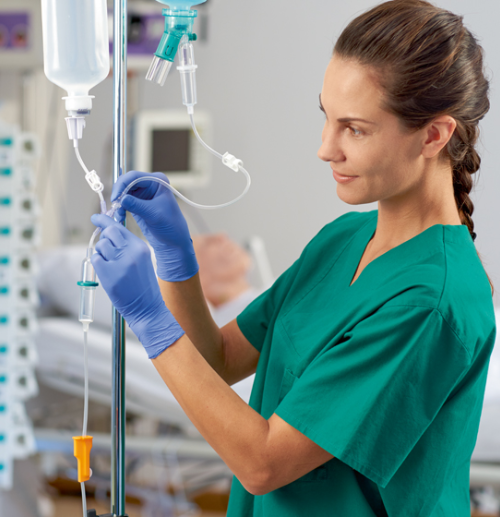 Ein medizinischer Mitarbeiter in grünem Kittel und blauen Handschuhen justiert eine Infusion. Die Szene scheint ein Krankenhauszimmer mit medizinischen Geräten zu sein, im Hintergrund liegt ein Patient in einem Bett.