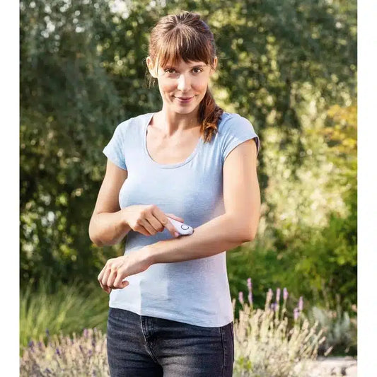 Eine Frau mit Pferdeschwanz, hellblauem T-Shirt und dunkler Jeans steht im Freien in einem Garten. Sie behandelt ihren Arm mit dem Gerät Beurer Insektenstichheiler BR 10 der Beurer GmbH. Im Hintergrund sind Grün- und Blütenpflanzen zu sehen.