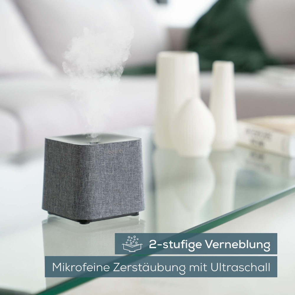 Een Beurer LA 35 aroma-diffuser van Beurer GmbH, vrij in grijs, siert een glazen tafel in een met meubels ingericht woonkamer. De tekst op de afbeelding luidt '2-staps verneveling' en 'Microfijne verstuiving met ultrasoon', wat de tweestaps verneveling en ultrafijne verstuiving door middel van ultrasone technologie benadrukt. Dit veelzijdige apparaat fungeert zowel als luchtbevochtiger als aroma-diffuser.