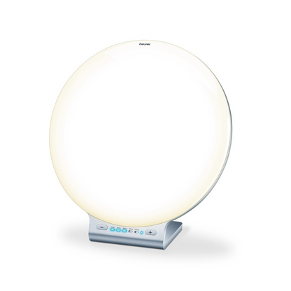 Een ronde, witte Beurer daglichtlamp TL 100 van Beurer GmbH op een zilveren standaard met een klein bedieningspaneel aan de onderkant met knoppen en een digitaal display. De lamp straalt een zacht, gelijkmatig licht uit, perfect als sfeerverlichting, met de merknaam „Beurer“ boven in het midden gedrukt.