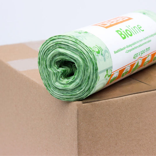 Un rouleau de sacs biodégradables verts est posé sur un carton. L'étiquette sur le rouleau est partiellement visible et porte l'inscription « EMIL DEISS KG (GmbH + Co.) ». Ces sacs 100 % compostables ont un design floral et mesurent 430 x 500 mm.
